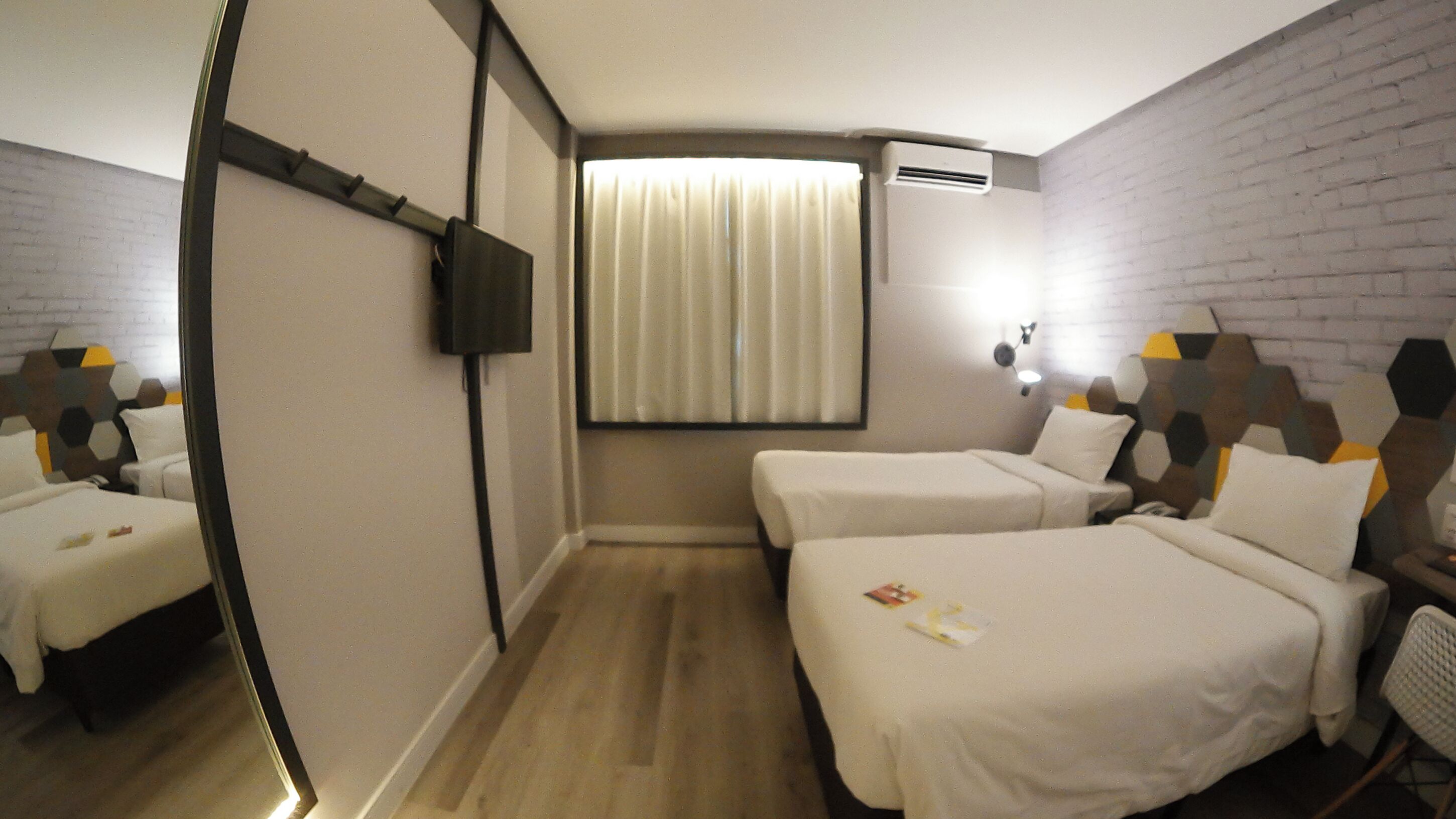Quarto de hotel com duas camas de solteiro, uma mesinha, uma cadeira, televisão e janela com cortinas.