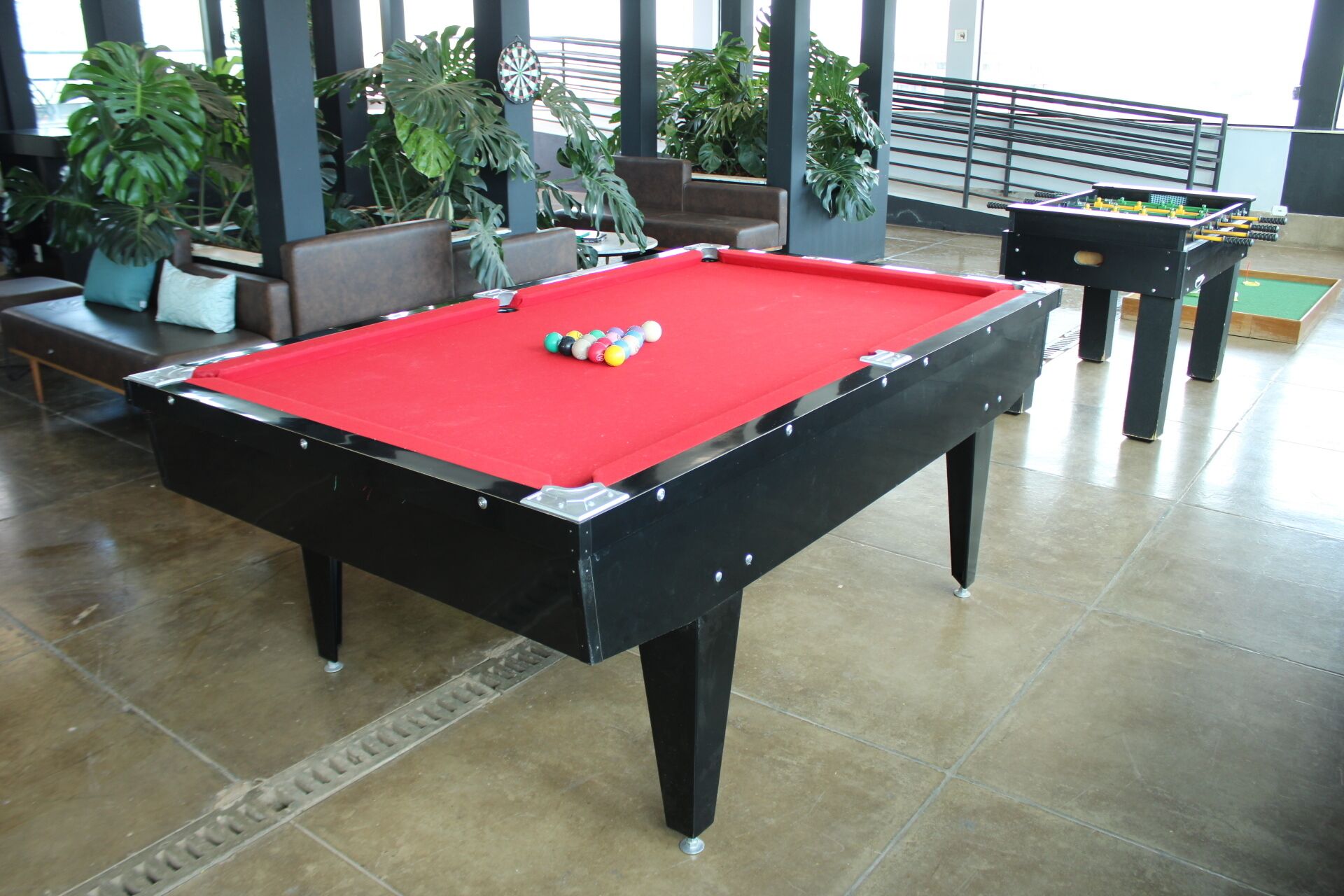 Une table de billard avec des billes, dans une pièce dont le sol est recouvert de carreaux et où l’on trouve des plantes en pot.