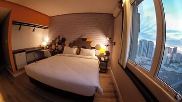 Quarto de hotel com uma cama grande, uma escrivaninha, uma cadeira e vista para a cidade através de uma janela ampla.