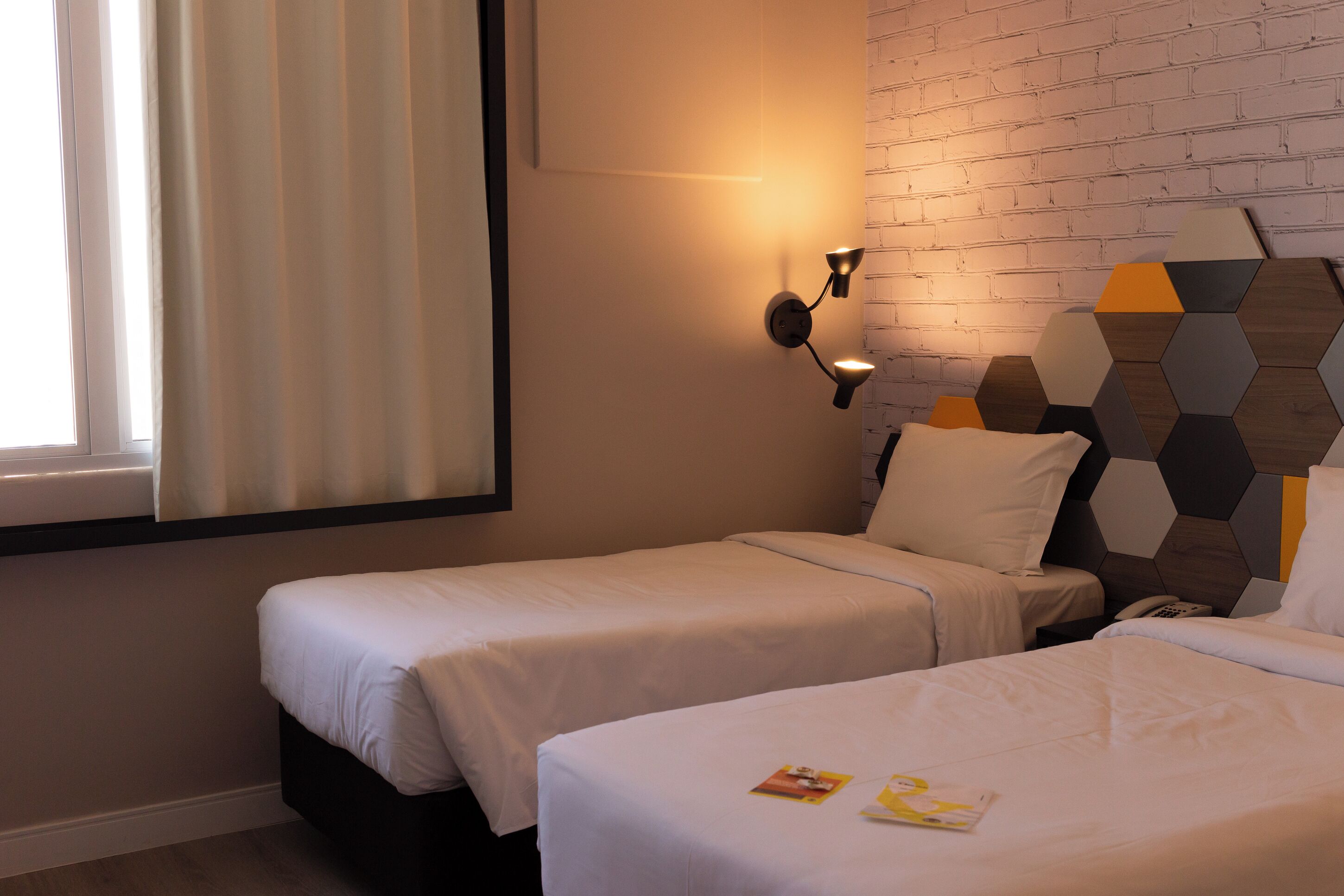 Habitación de hotel con dos camas individuales, una lámpara de pared y un cabecero con motivos geométricos.