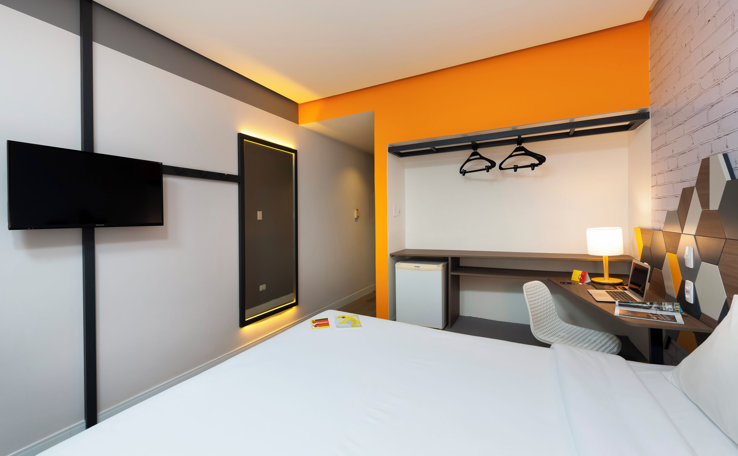 Une chambre d’hôtel comprenant un lit, un bureau avec une chaise, un téléviseur fixé au mur et un porte-manteau avec des cintres.