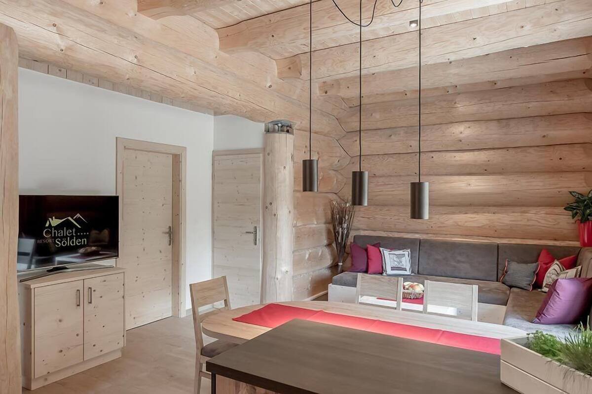 Un interior moderno con paneles de madera, una zona de comedor con camino de mesa rojo, un sofá con cojines y un televisor mostrando 'Chalet... RESORT SÖLDEN'.