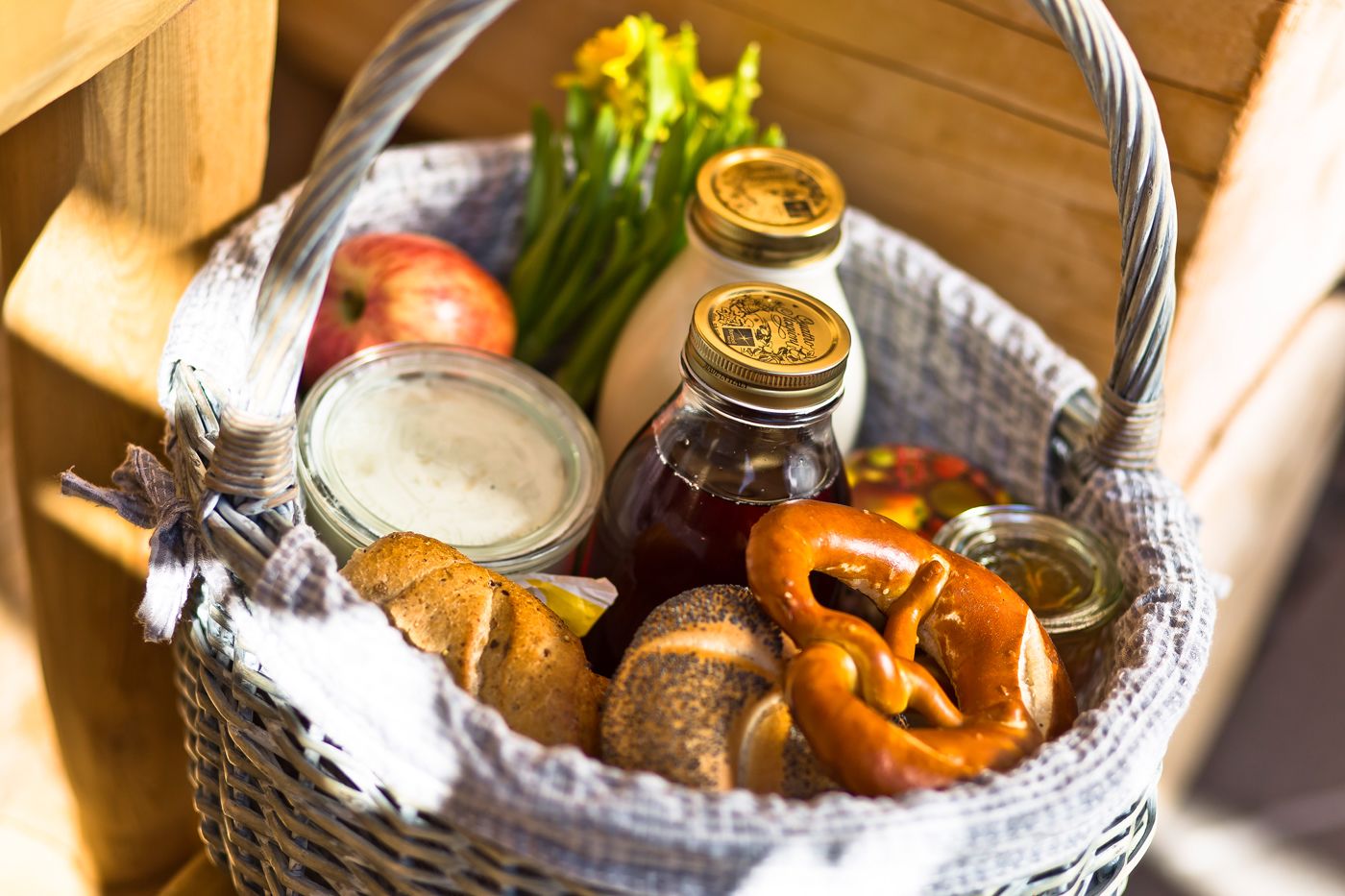 A wicker basket containing pretzels, a jar of mustard, a bottle of ketchup, a jar of mayonnaise, an apple, and some pickles.