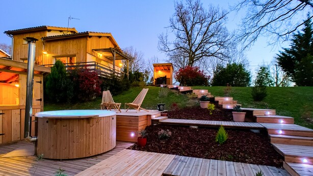 A wooden deck with a hot tub, wooden chairs, and a small building with lights. - Gîte dans les bois - Nordic SPA & Nature (Montdurausse)