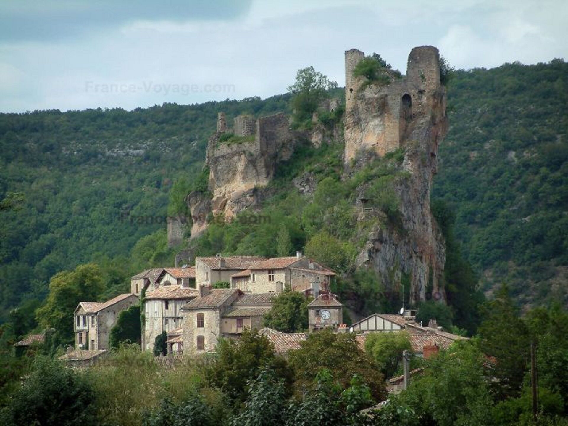 A historic stone building with a clock tower, surrounded by lush greenery and a rocky cliff.