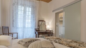 Un dormitorio con cama, una silla de madera, tocador, espejo y ventana con cortinas florales.