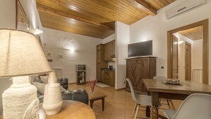 Un comedor con mesa de madera, sillas y una lámpara. Se ve una cocina al fondo con armarios y un televisor.