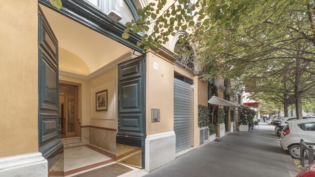 Entrada de hotel con puerta doble, un pequeño vestíbulo y vista a la calle con coches aparcados y un árbol.