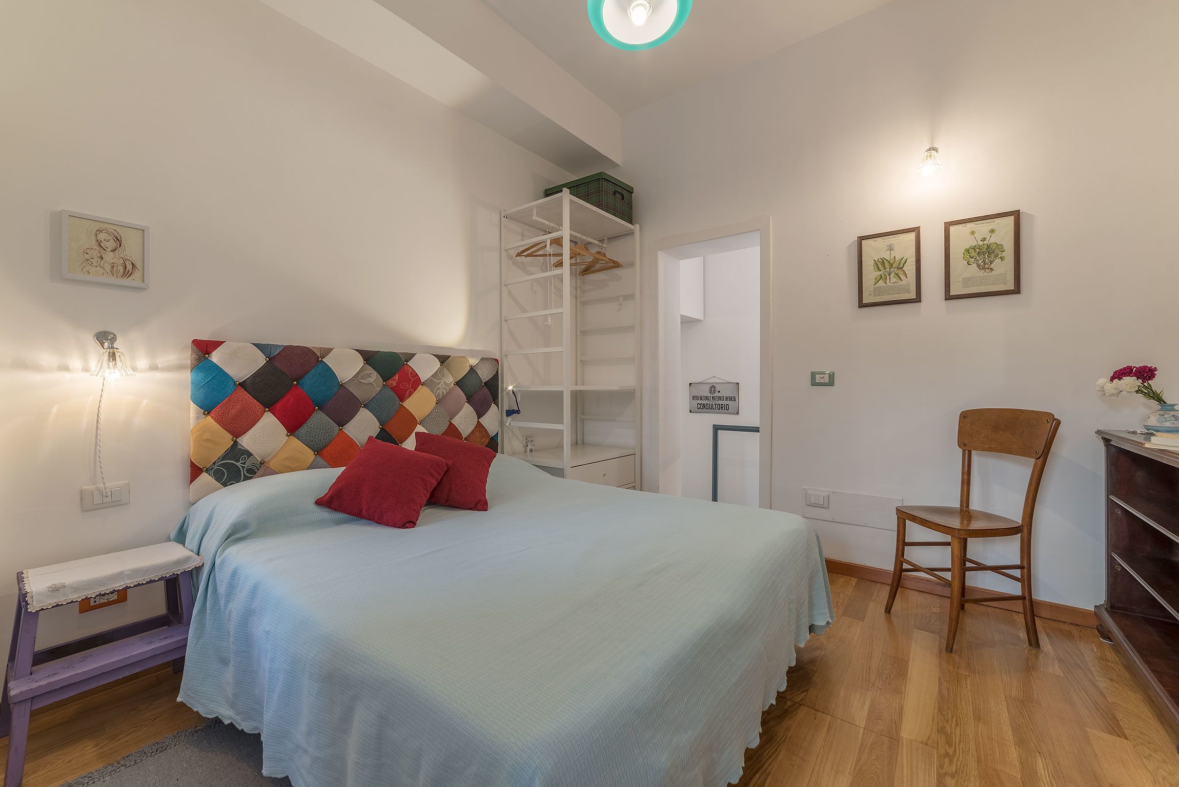 A bedroom with a bed, a bedside table, a chair, a wardrobe, and two framed botanical prints on the wall.