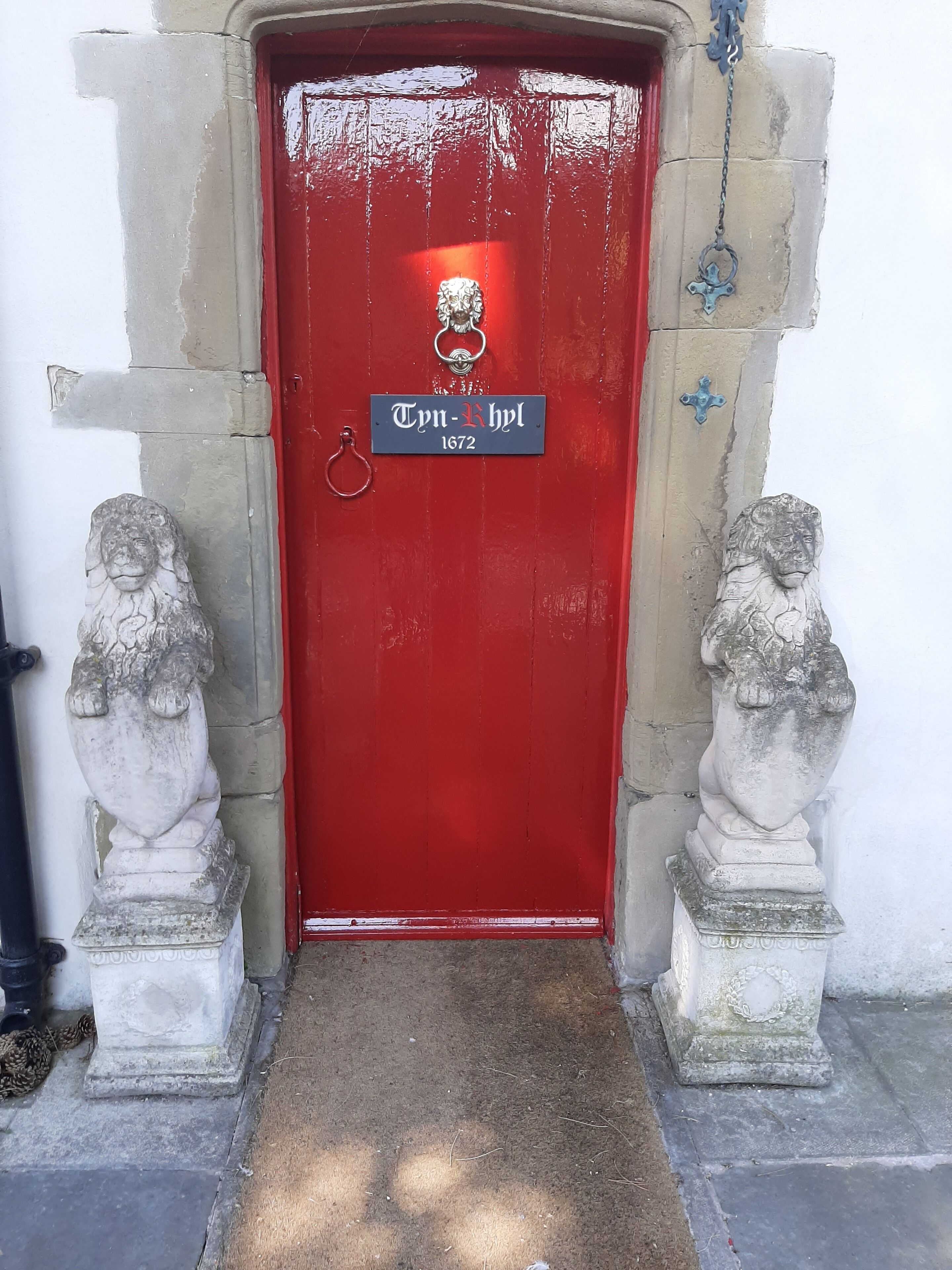 Une porte rouge ornée d’une plaque portant l’inscription « Tyn-Khyl 1672 » et flanquée de deux statues de lions en pierre, une de chaque côté.
