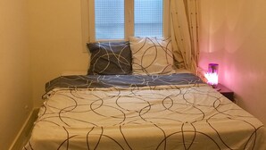 Une chambre à coucher avec un lit, une fenêtre avec des rideaux, une table de chevet avec une lampe et un plancher carrelé.