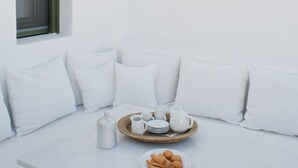 Ein weißes, minimalistisches Zimmer mit einer weißen Bank, Kissen, einer Korbtasse mit Tassen und Tellern sowie einem kleinen Tisch mit einer Karaffe.
