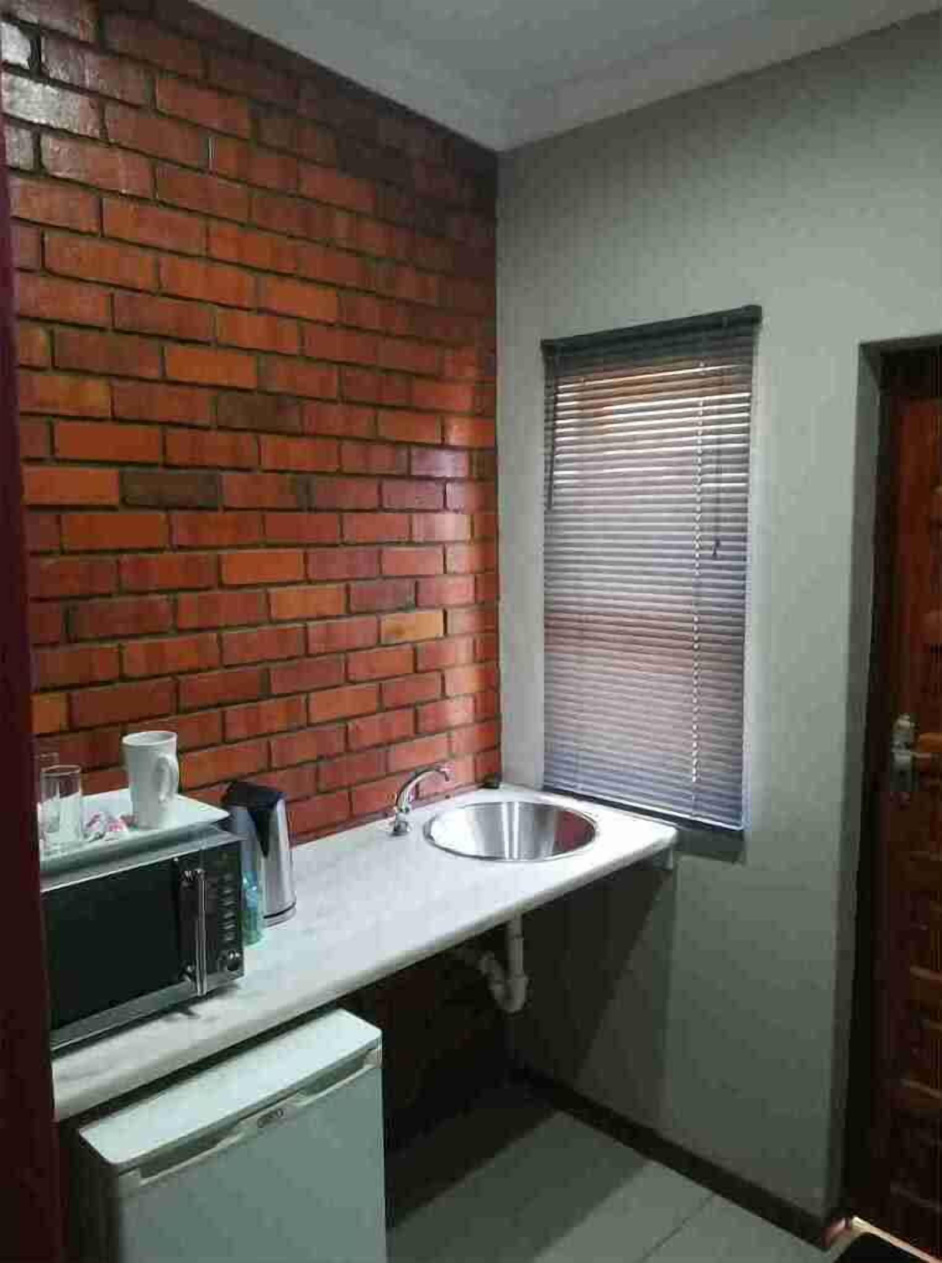 A compact kitchen with a brick wall, a sink, a window with blinds, and a microwave.