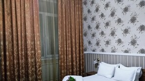 Un lit bien fait, avec du linge de lit blanc et des oreillers, une petite table de chevet avec une lampe, et une fenêtre ornée de rideaux à motifs.