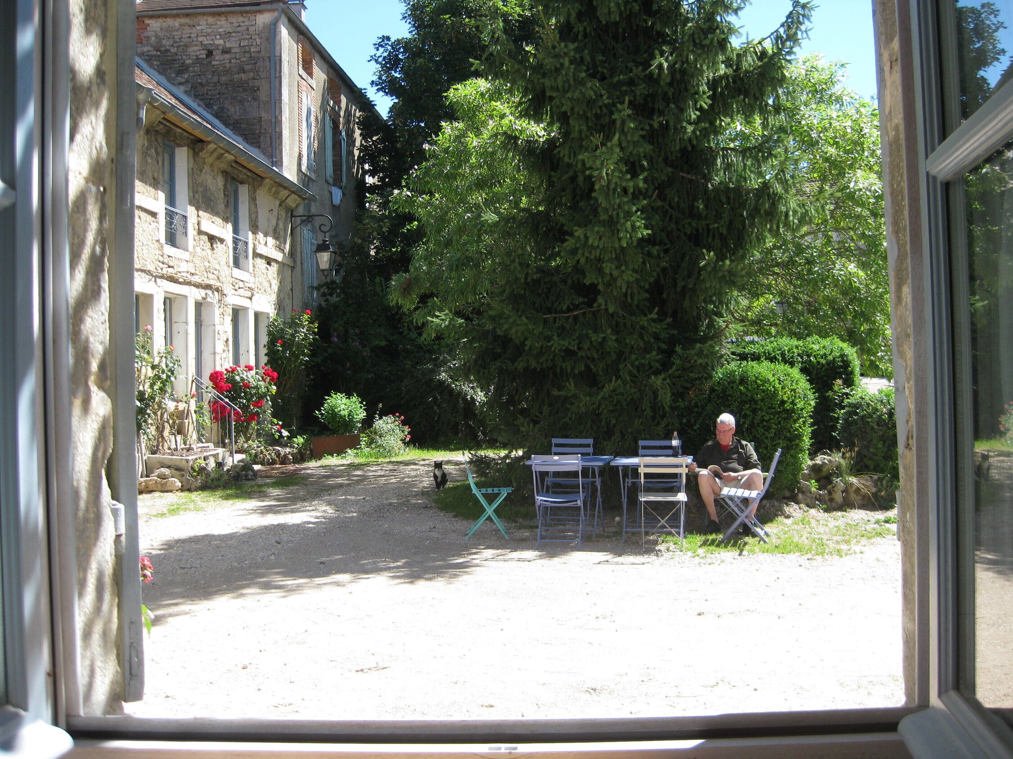 A courtyard with a gravel path, a large tree, and a building with a balcony. There are outdoor chairs and a table.