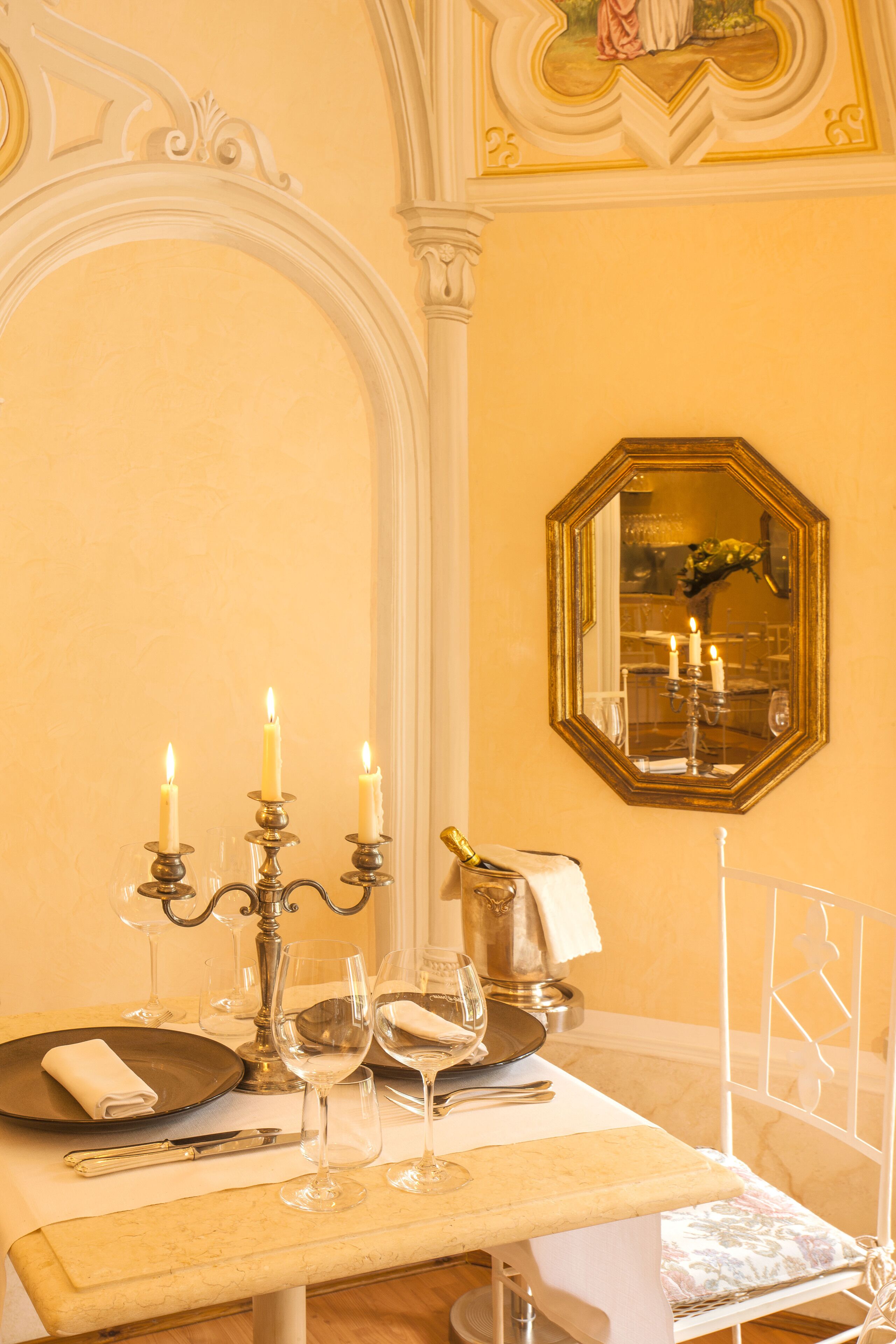 Una mesa de comedor dispuesta con velas, copas y cubiertos, con un espejo y detalles ornamentales en el techo.