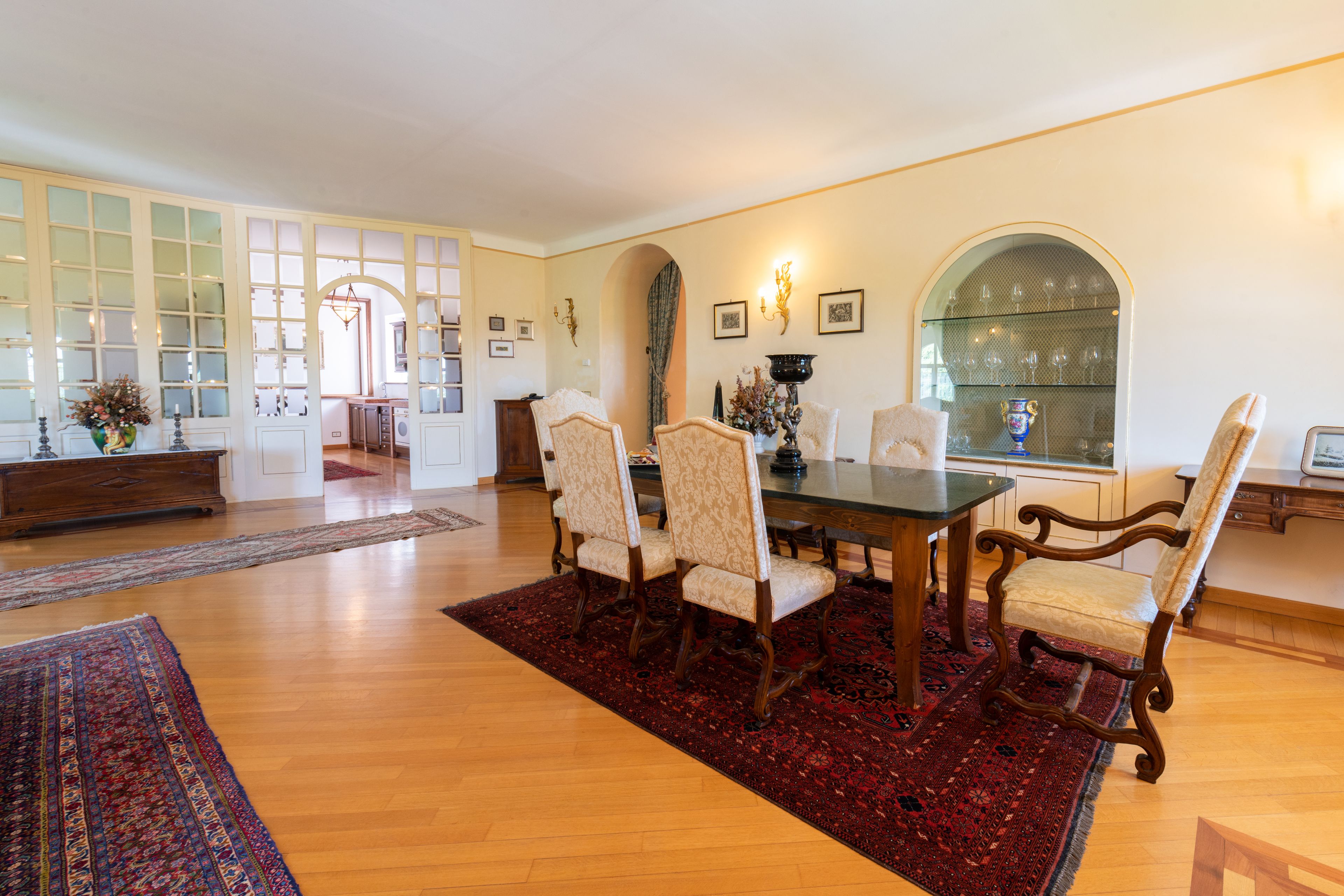 A dining area with a wooden table and chairs, a red rug, and a view of the kitchen through an archway.