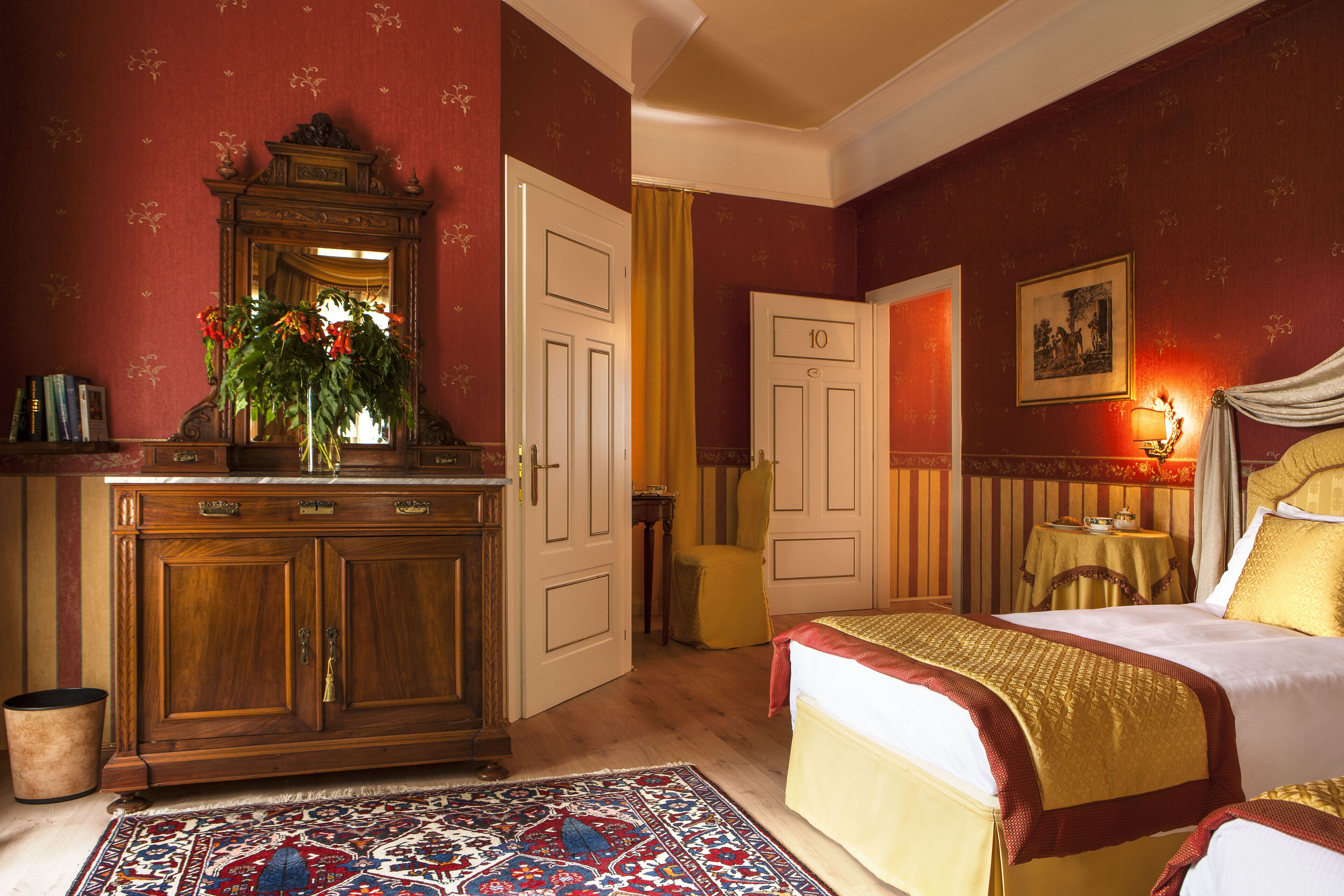 A traditional room with a wooden dresser, a bed with a red and gold bedspread, a mirror with a floral arrangement, and a small table with a lamp.
