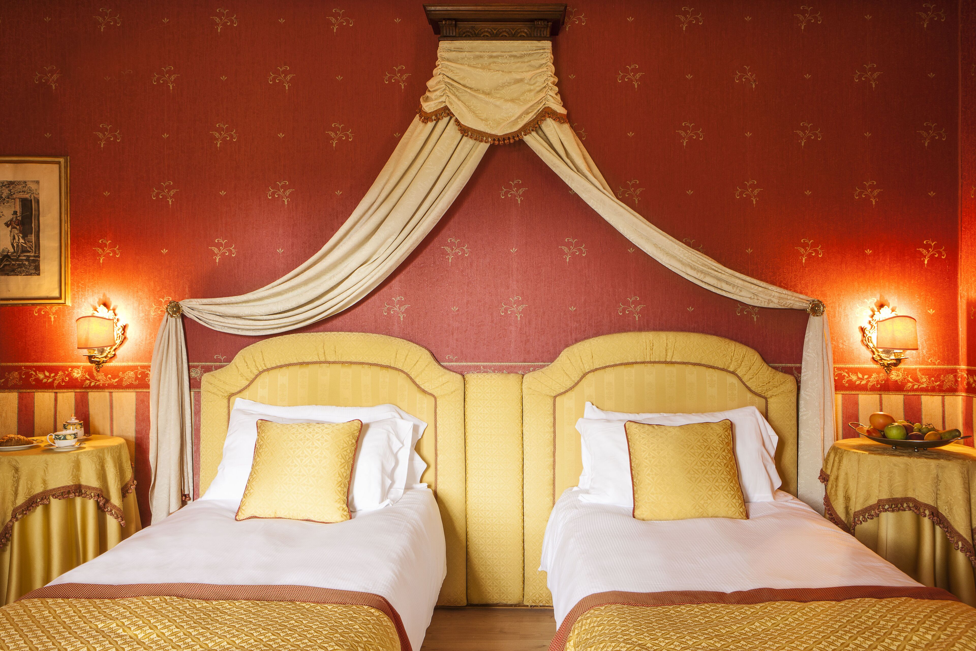 Une chambre d’hôtel avec deux lits, un baldaquin et un panier de fruits posé sur une table d’appoint.