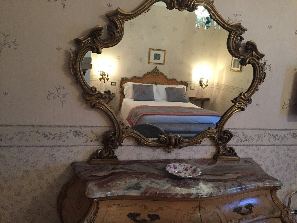 A bedroom with a large ornate mirror, a bed with a floral bedspread, and a dresser with a decorative plate.