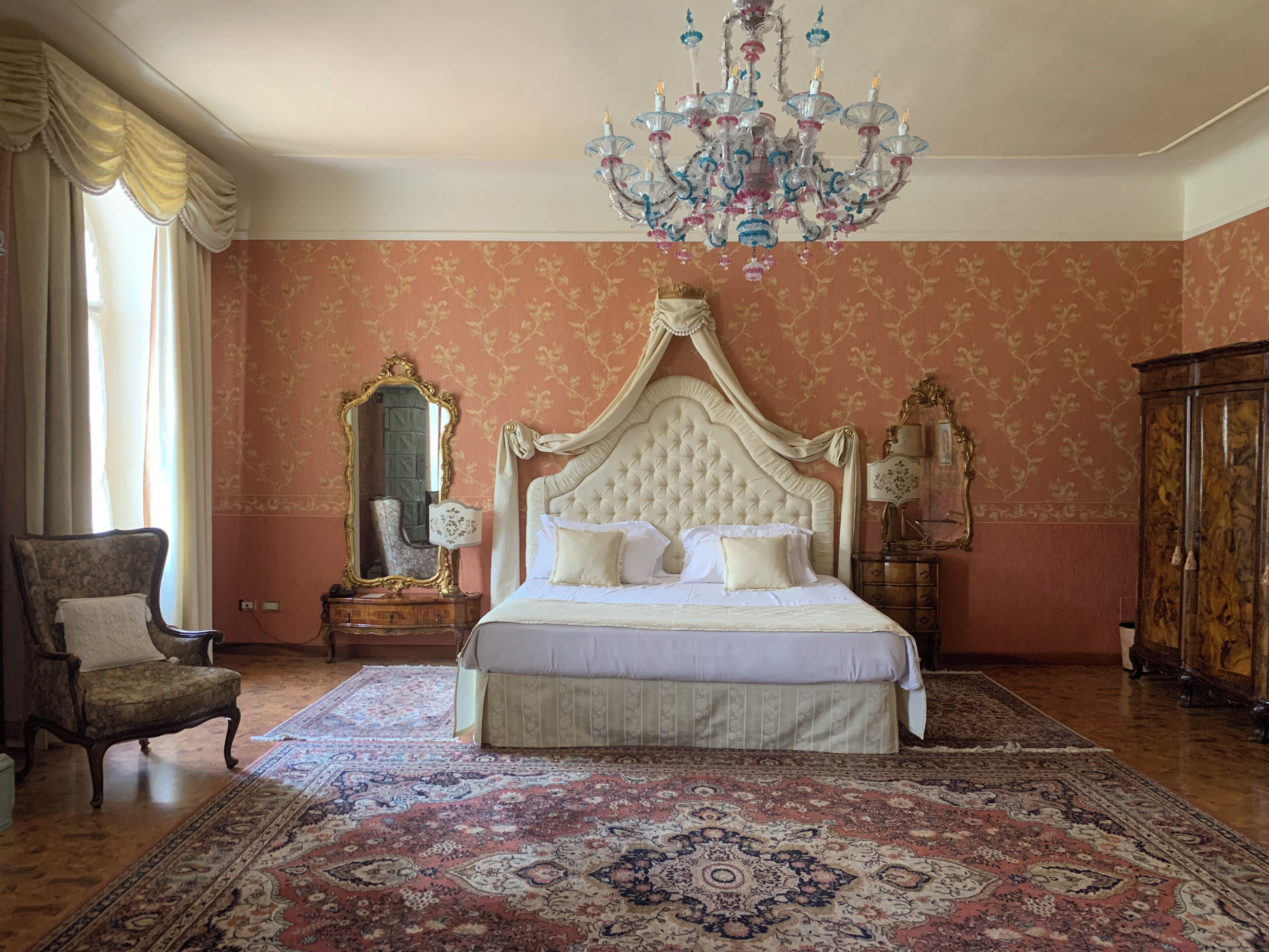 A bedroom with a large bed, a chandelier, a mirror, a chair, and a cabinet.