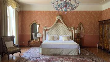 A bedroom with a large bed, a chandelier, a mirror, a chair, and a cabinet.