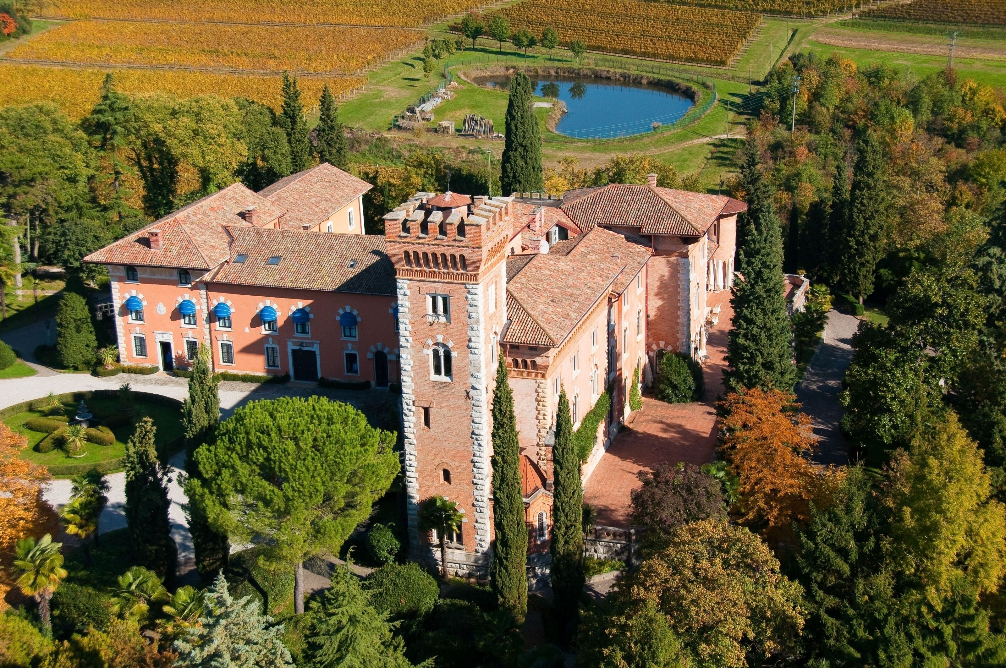 A large, historic building with a central tower and multiple wings, surrounded by lush gardens and a pond.