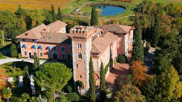 A large, historic building with a central tower and multiple wings, surrounded by lush gardens and a pond.