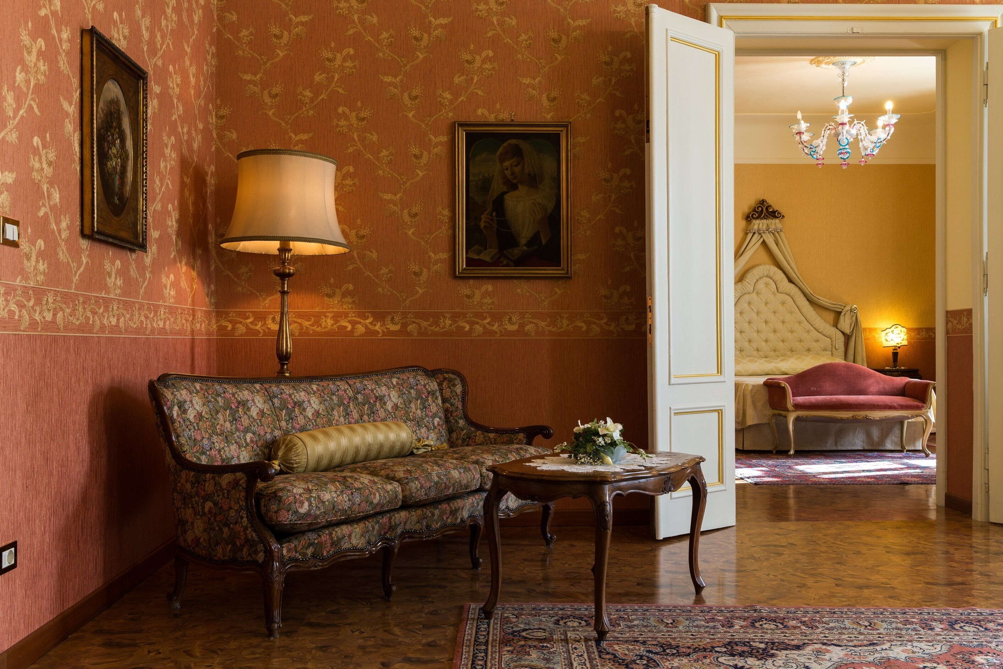 A room with a floral-patterned sofa, a table with a vase of flowers, a bed with a canopy, and a chandelier.