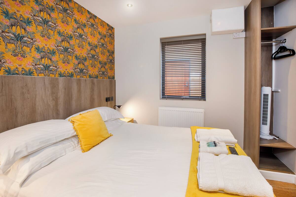 Habitación de hotel con una cama, una manta amarilla, una toalla blanca, una ventana con persianas, un radiador y un armario.