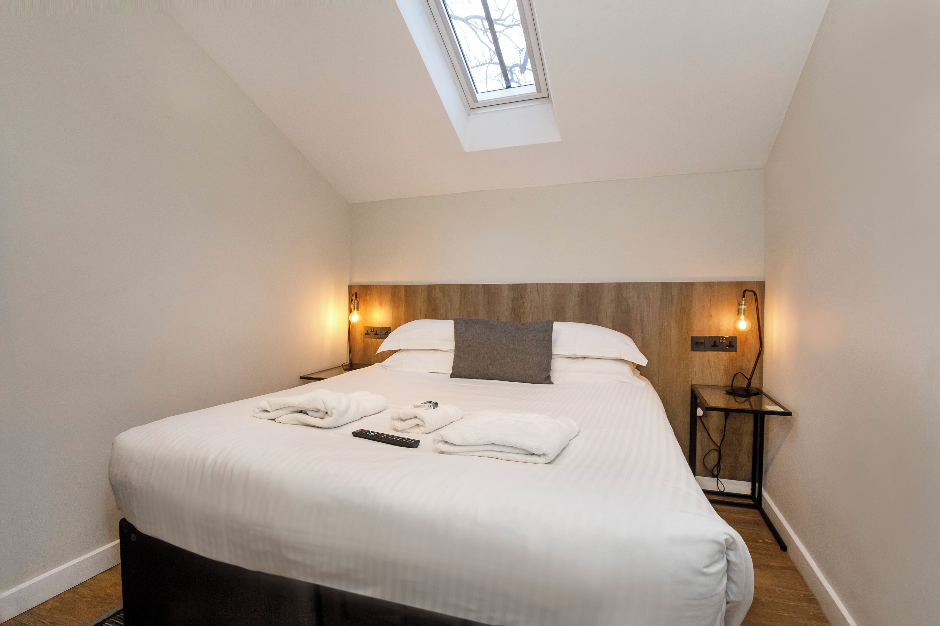 Een net opgemaakt bed met witte lakens, een grijs kussen, en twee handdoeken die op het bed gevouwen zijn. Er staat een klein nachtkastje met een lamp en een stopcontact. De kamer heeft een schuine kap met een dakraam, houten lambrisering aan de muur en een witte deur.