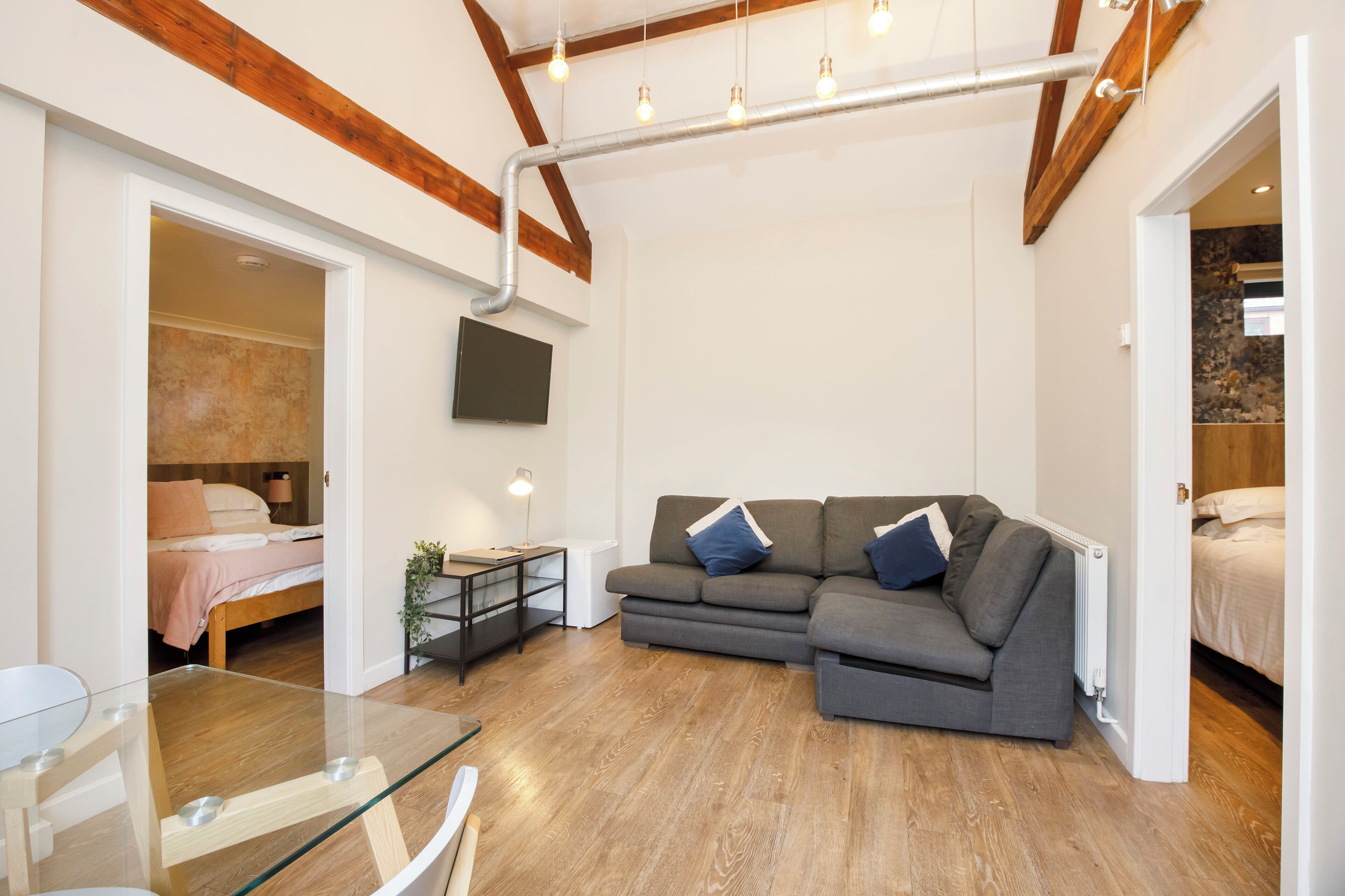 A modern living room with a glass table, a grey sofa with blue cushions, wooden beams on the ceiling, and a flat-screen TV mounted on the wall.