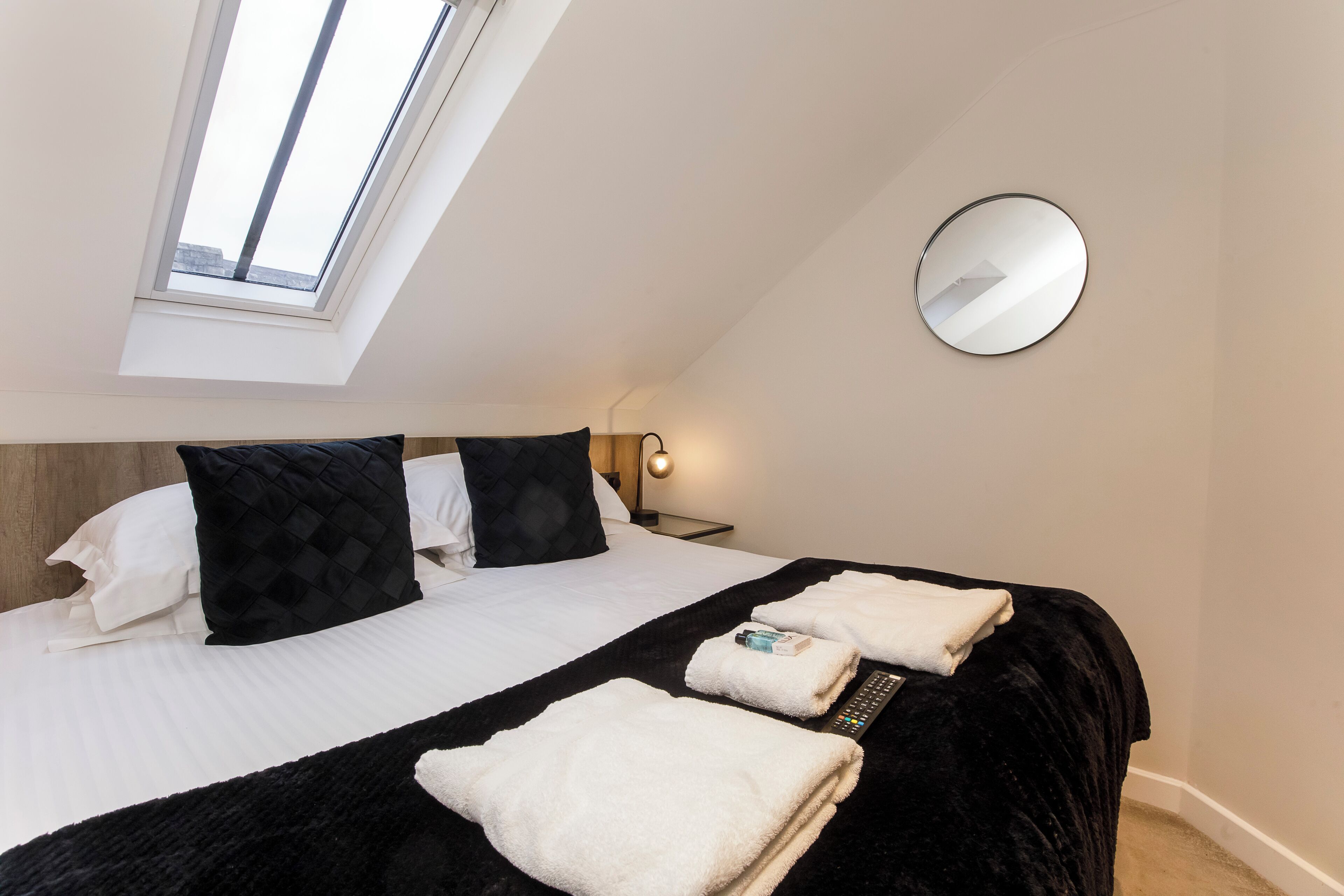 A bedroom with a skylight, a round mirror, a bed with white and black bedding, and a remote control on the bed.