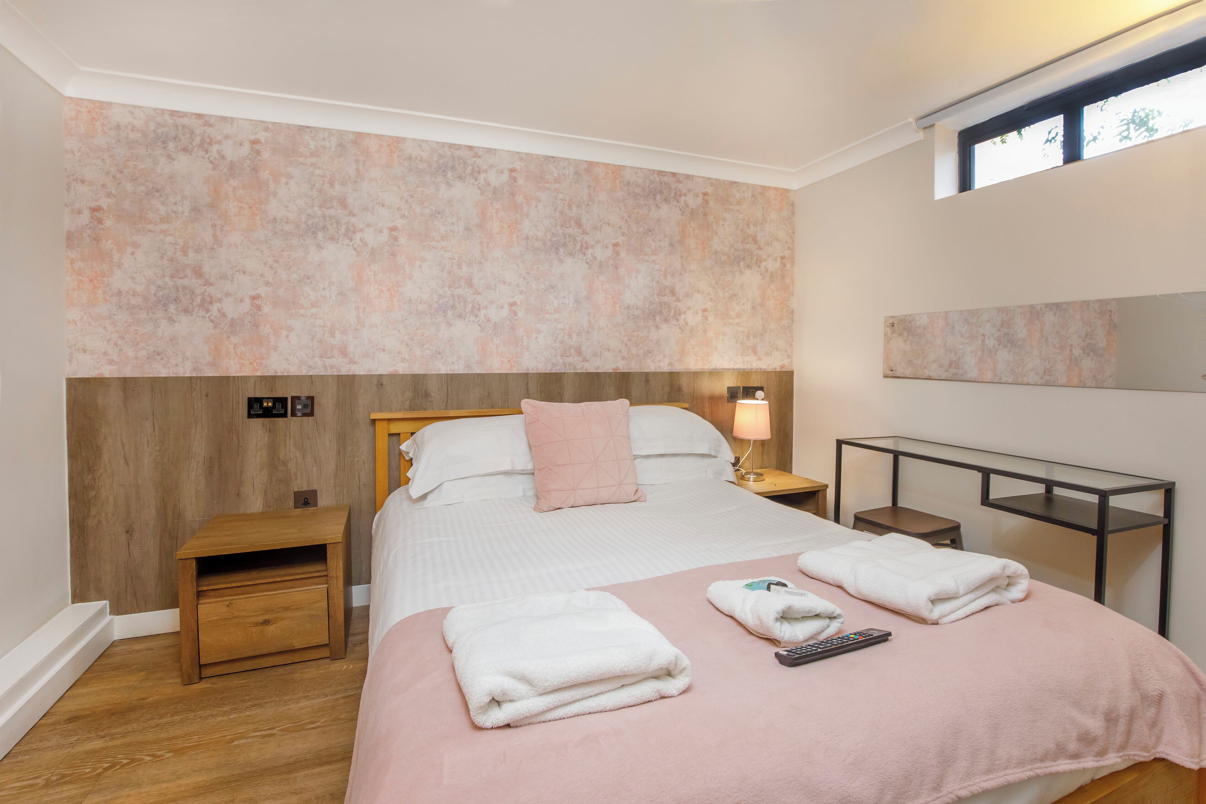 Ein ordentlich bezogenes Bett mit einer rosa Decke, ein Nachttisch aus Holz, ein kleiner Tisch und ein an der Wand befestigtes Regal.