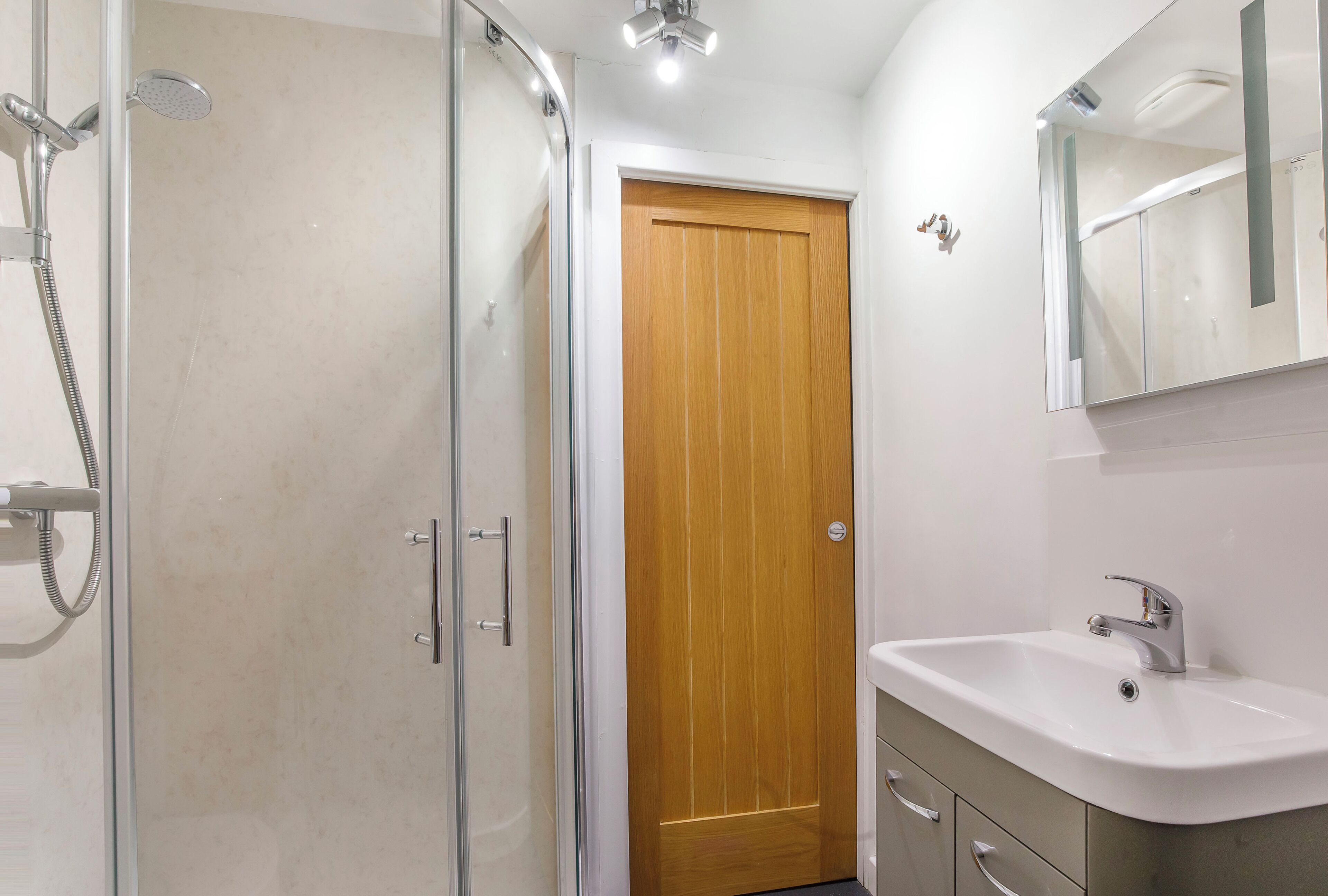 A bathroom with a glass shower enclosure, a wooden door, a sink with a mirror, and a cabinet with drawers.