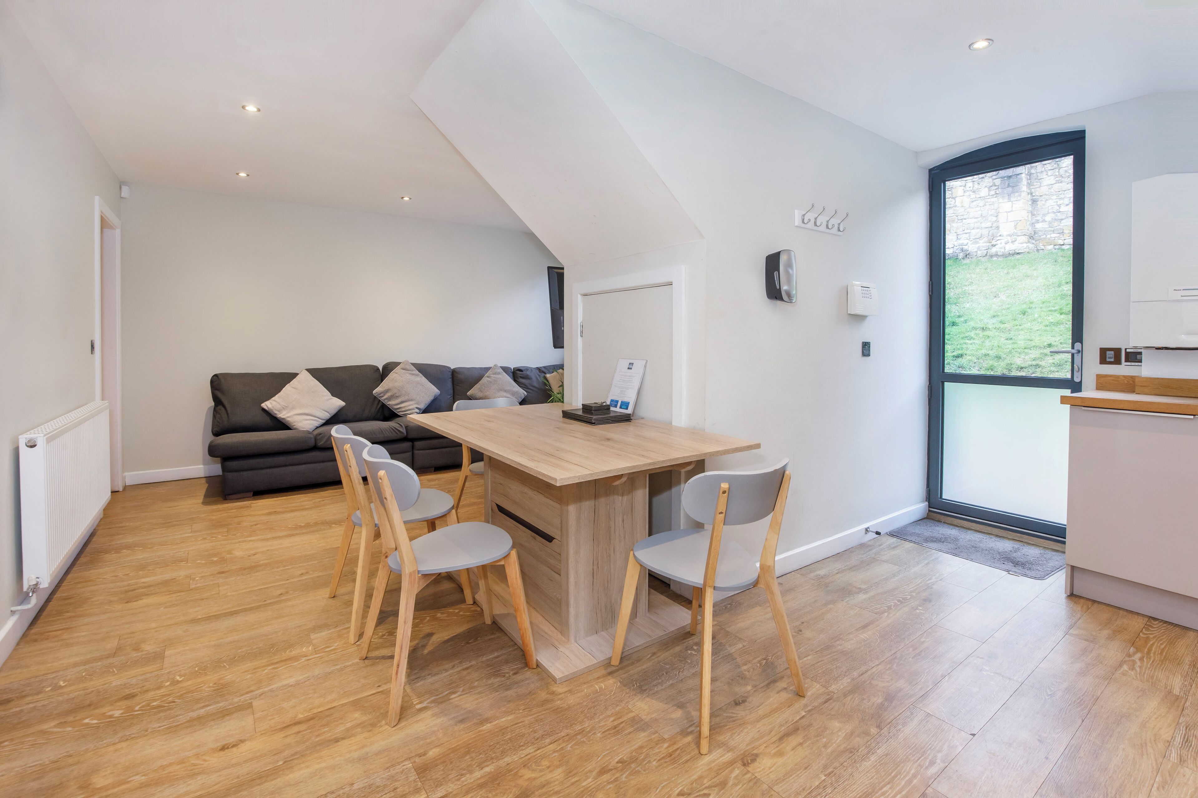 A modern kitchen with a wooden dining table and chairs, a grey sofa, and a large window.