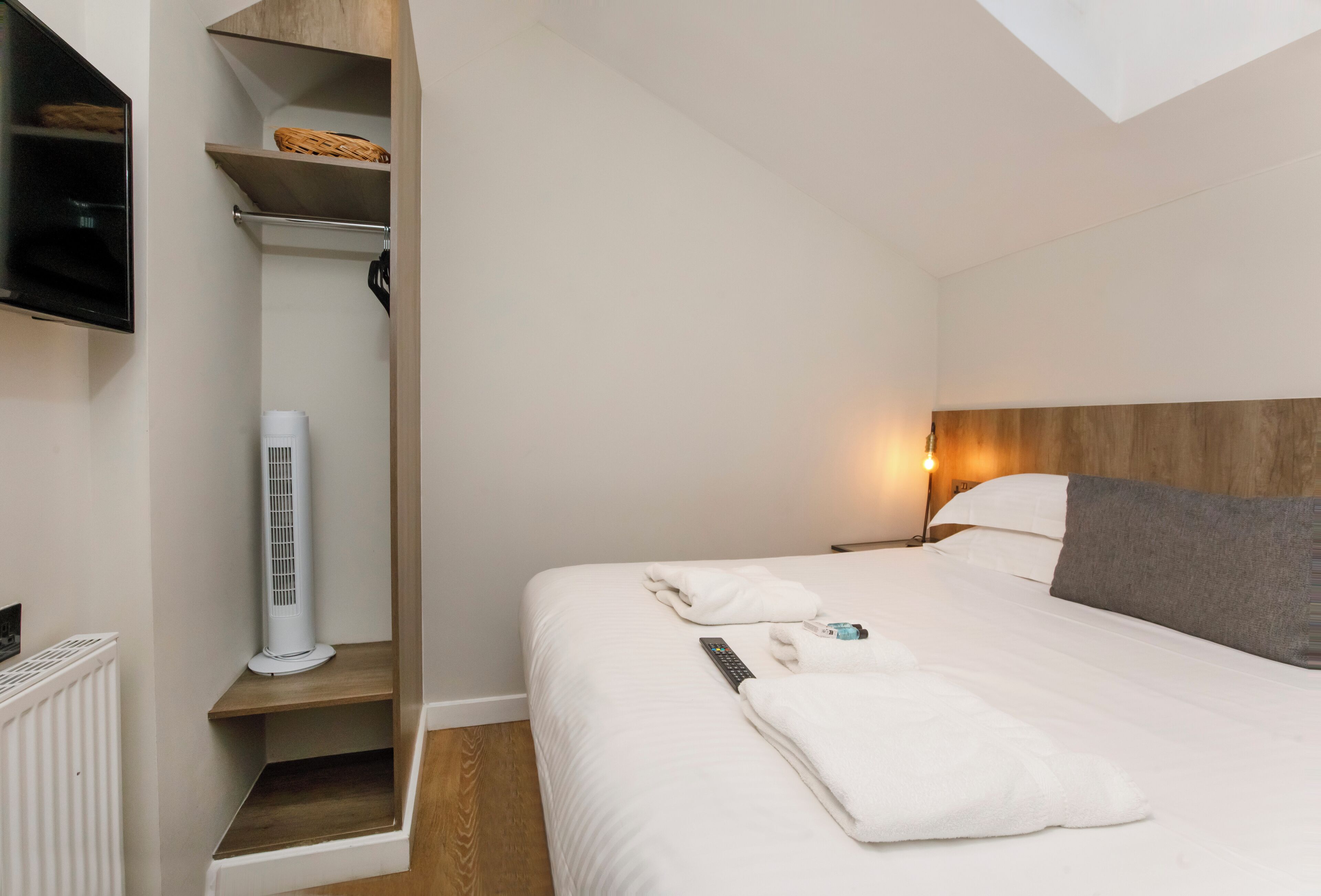 A neatly made bed with white linens, a remote control, and a towel on the bed. There is a white air conditioner on a shelf, and a wooden headboard with a light fixture above the bed.