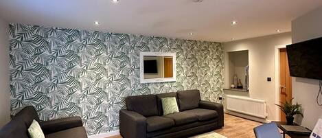 A modern living room with a patterned wallpaper, two sofas, a coffee table, and a TV mounted on the wall.