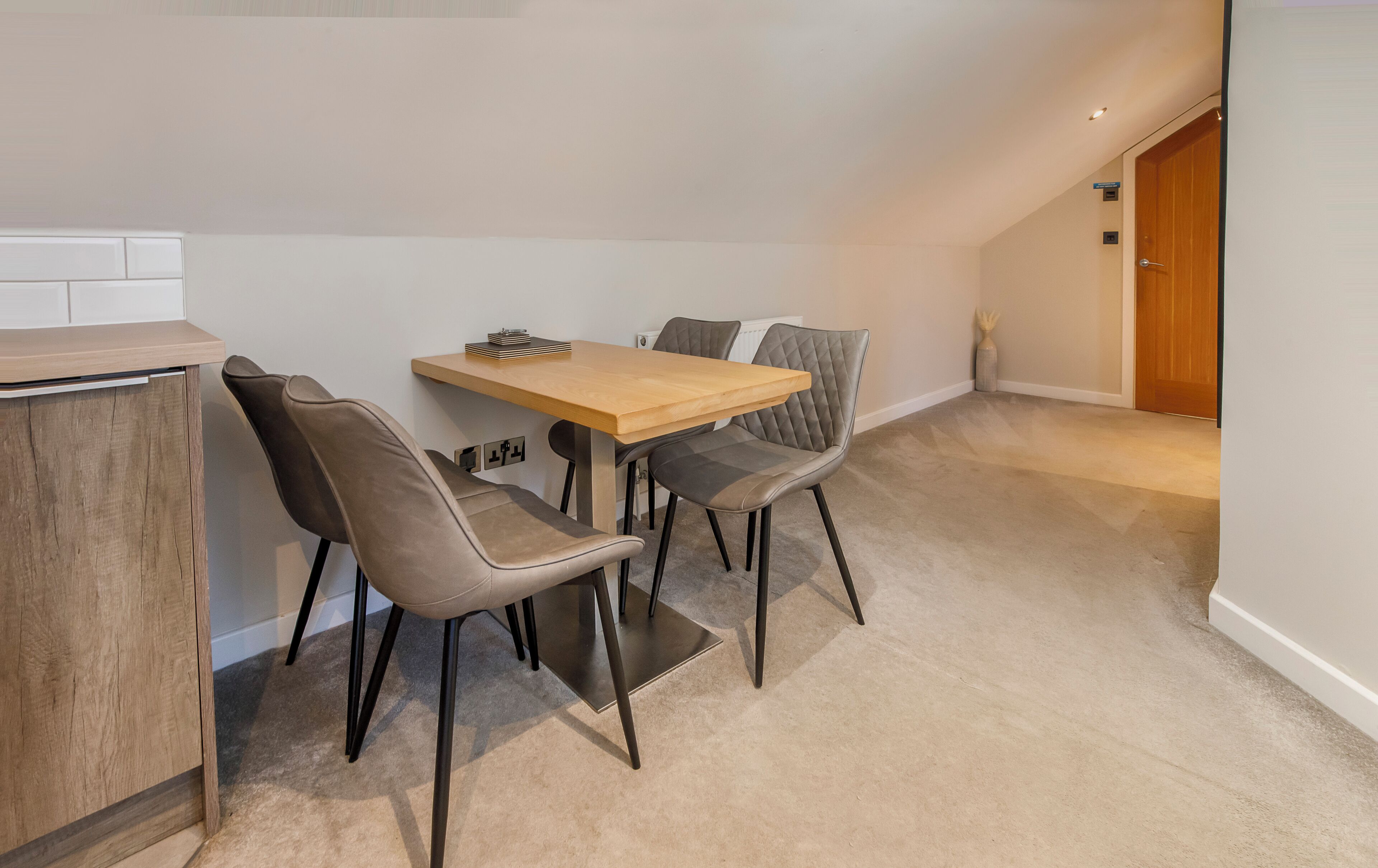 A small, modern room with a wooden dining table and two chairs, a radiator, and a door leading to another room.