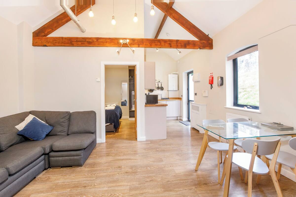 Una sala moderna con un sofá modular, una mesa de comedor de vidrio y sillas de madera. Se ve una cocina al fondo con armarios blancos y una ventana.