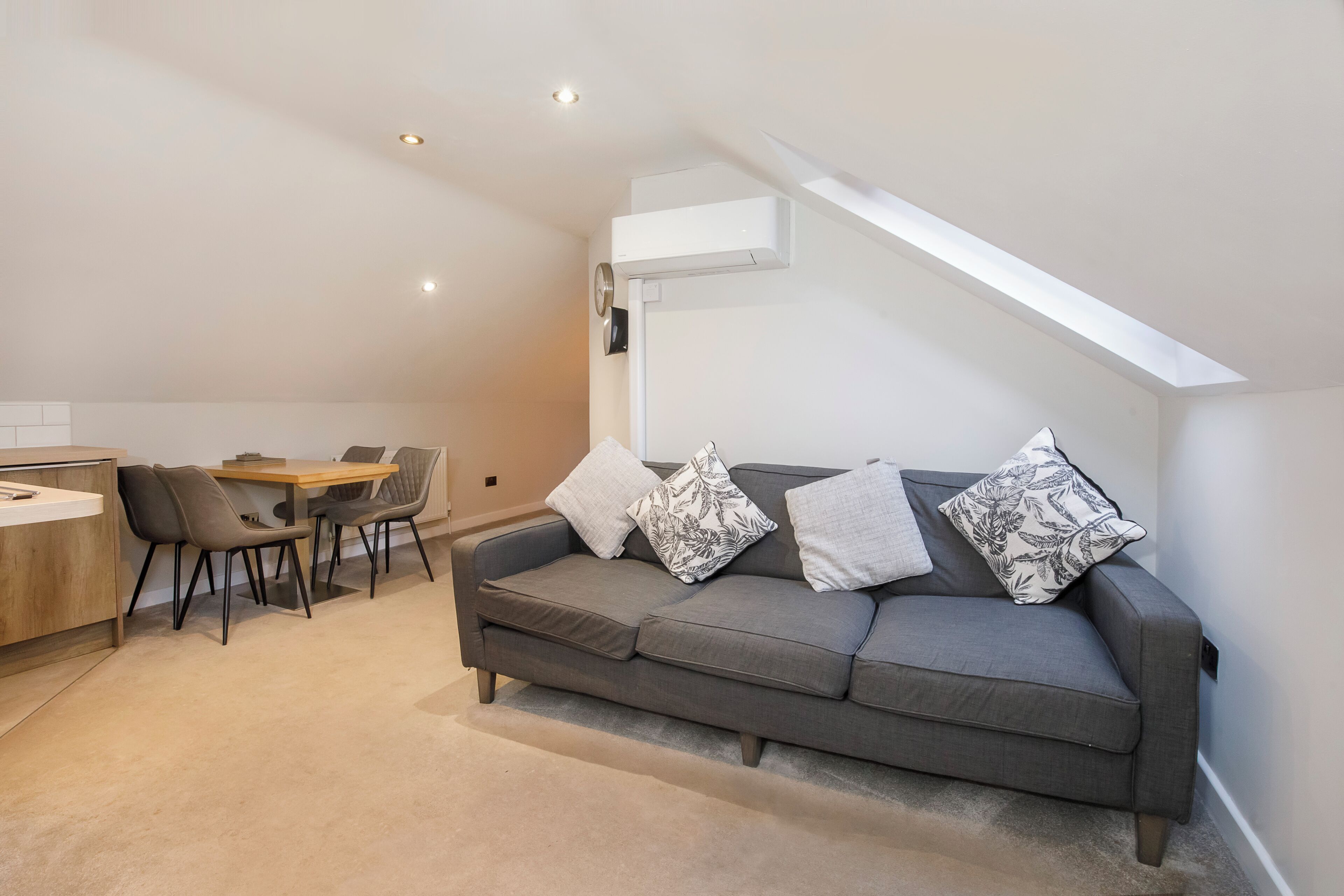 A modern loft apartment with a gray sofa, two chairs, a small wooden table, and a built-in kitchen counter.