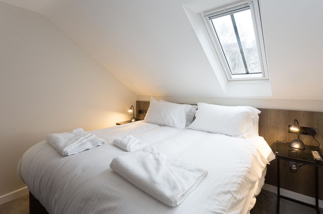 A neatly made bed with white linens and towels, a bedside table with a lamp, and a window with a view of trees.