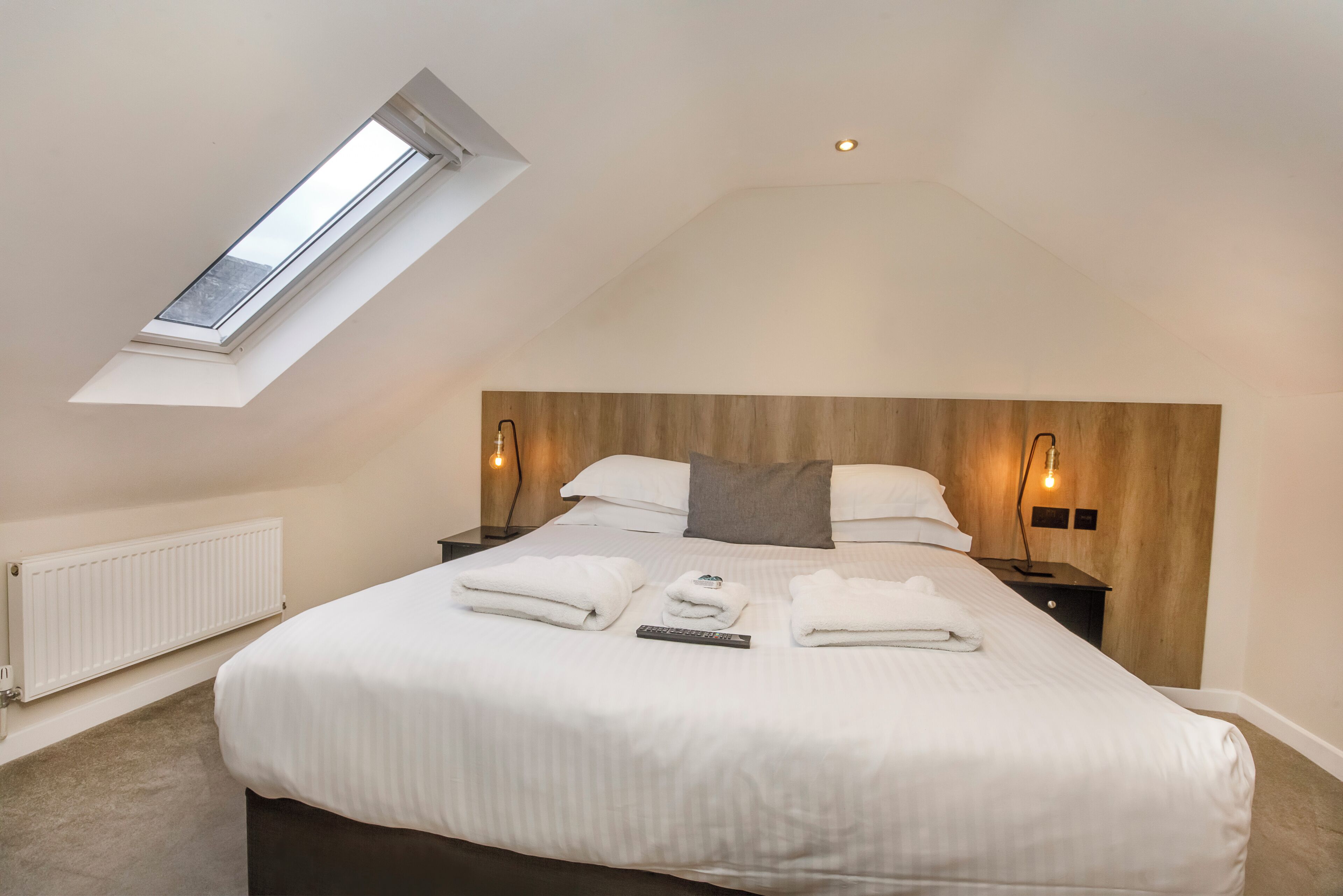 A bedroom with a skylight, a bed with white linens, two bedside tables with lamps, and a wooden headboard.