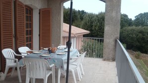 Una veranda esterna con tavolo e sedie, un ombrellone grande e vista su una casa e degli alberi.