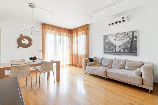 A modern living room with a wooden floor, a beige sofa, a dining table with chairs, a chandelier, and a large window with sheer curtains. - Tedeschi (Padova)