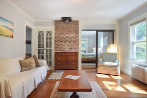 Une pièce avec une cheminée en briques, un canapé, une table en bois et une vue sur une chambre à coucher, visible à travers une porte vitrée.