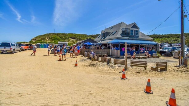 Un bâtiment situé au bord de la plage, doté d’un toit en pente, avec une plage de sable devant et des véhicules garés.
