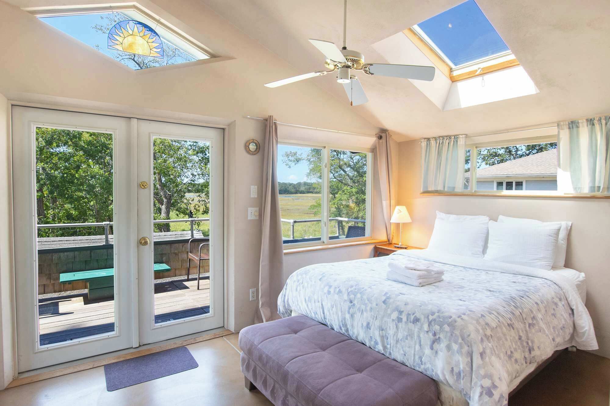 A bedroom with a bed, a ceiling fan, a skylight, and a view of a deck and outdoor area.