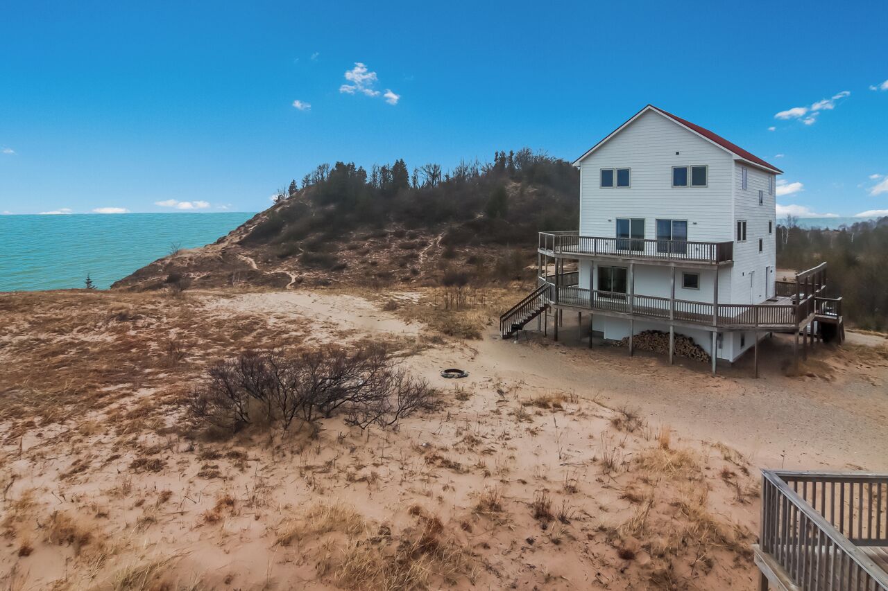 A two-story beach house with a balcony, situated on a sandy beach with a view of the ocean.
