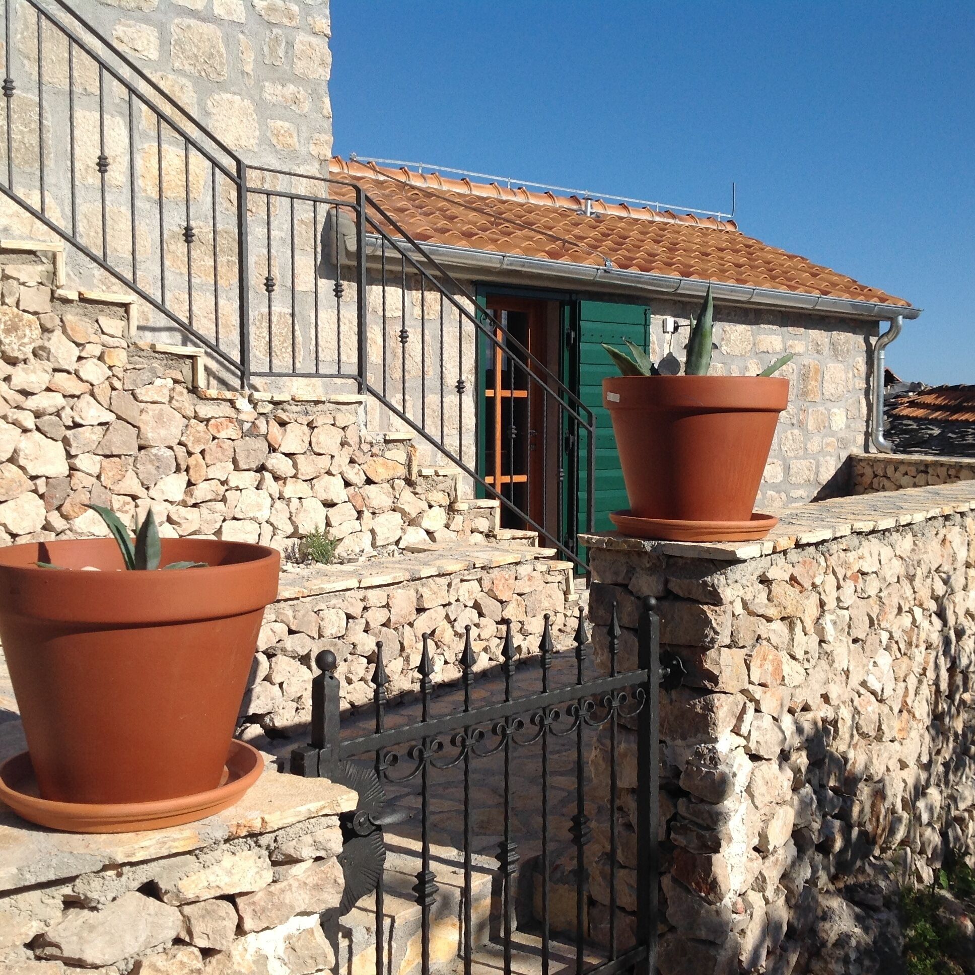 Un edificio de piedra con una escalera que lleva a una puerta verde, grandes macetas de terracota y una barandilla de metal.